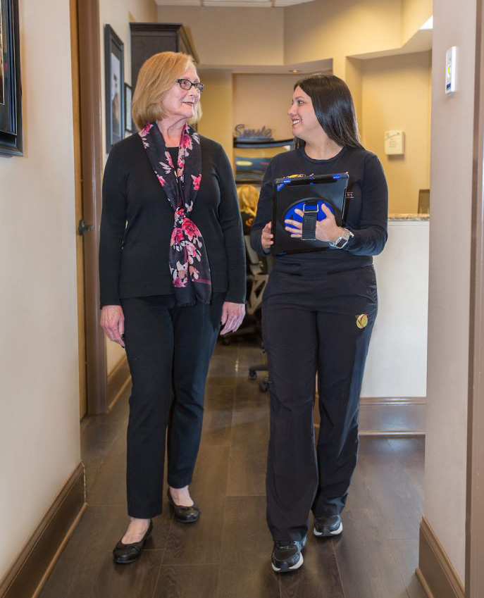 The image shows two individuals standing in an indoor setting with a professional appearance, likely a healthcare facility, where one person appears to be holding a clipboard and the other is wearing a badge, possibly indicating roles within their work environment.