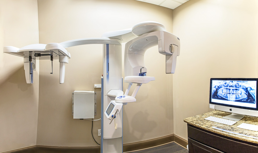 The image shows a dental office with a modern chair and equipment, including an overhead camera system, and a monitor displaying a patient s mouth.