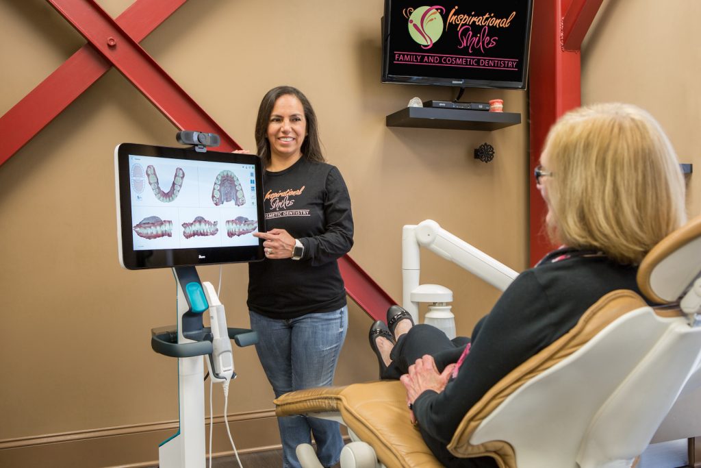 A woman standing next to a dental chair, smiling at the camera, with a television displaying teeth models behind her.