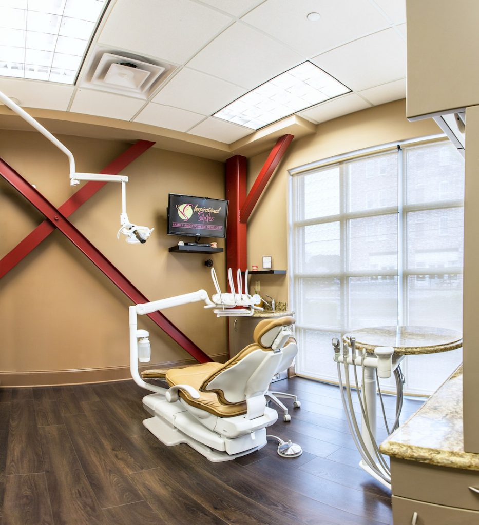 The image shows an interior view of a dental office with modern equipment and fixtures, including a large chair for dental procedures, a dental station with multiple chairs, and various dental tools and instruments. The room has a clean and professional appearance with a color scheme that includes shades of brown, red, and beige.