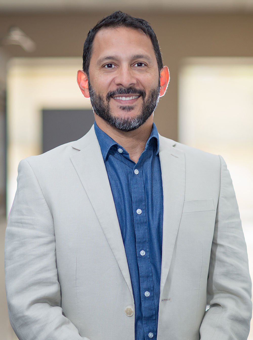 A man wearing a blue shirt and glasses stands smiling against a beige background.
