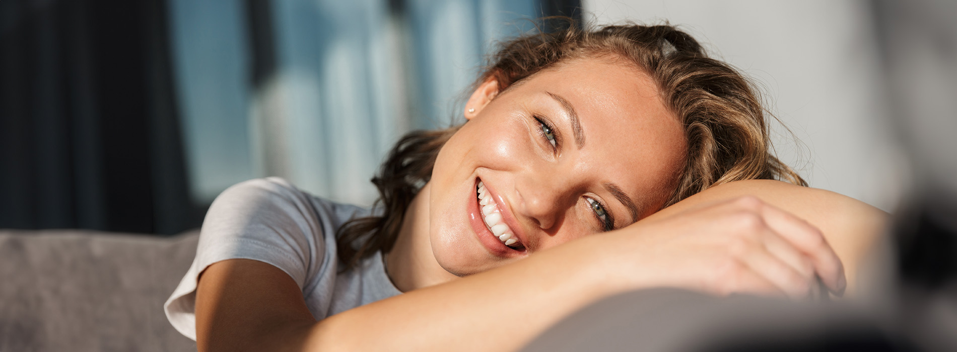 A woman with blonde hair laying on her side on a couch, smiling and looking towards the camera.