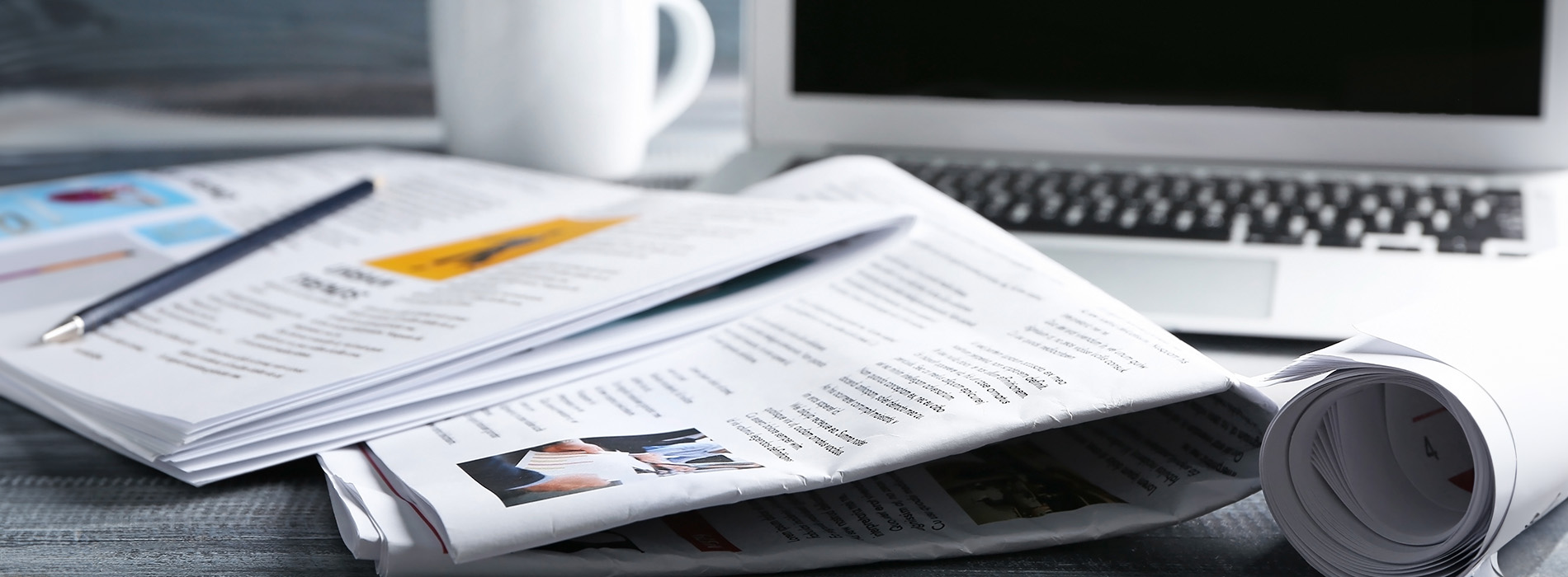 The image shows a collection of open newspapers spread out on a table with a laptop computer and a cup nearby, suggesting a workspace environment.