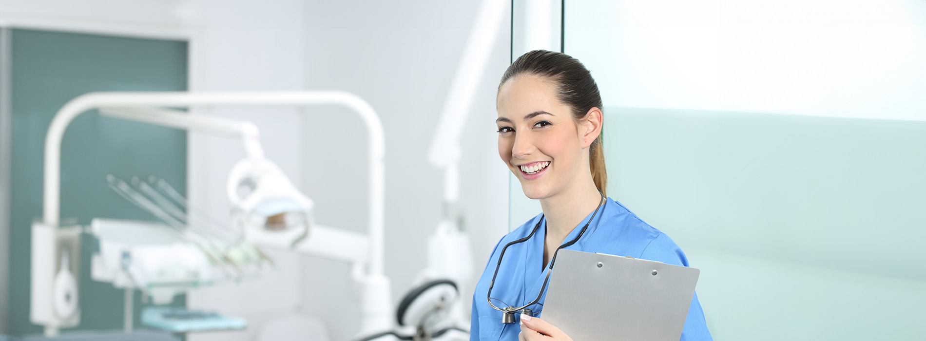 A female dental hygienist wearing blue scrubs, holding a clipboard, standing in a well-equipped dental office.