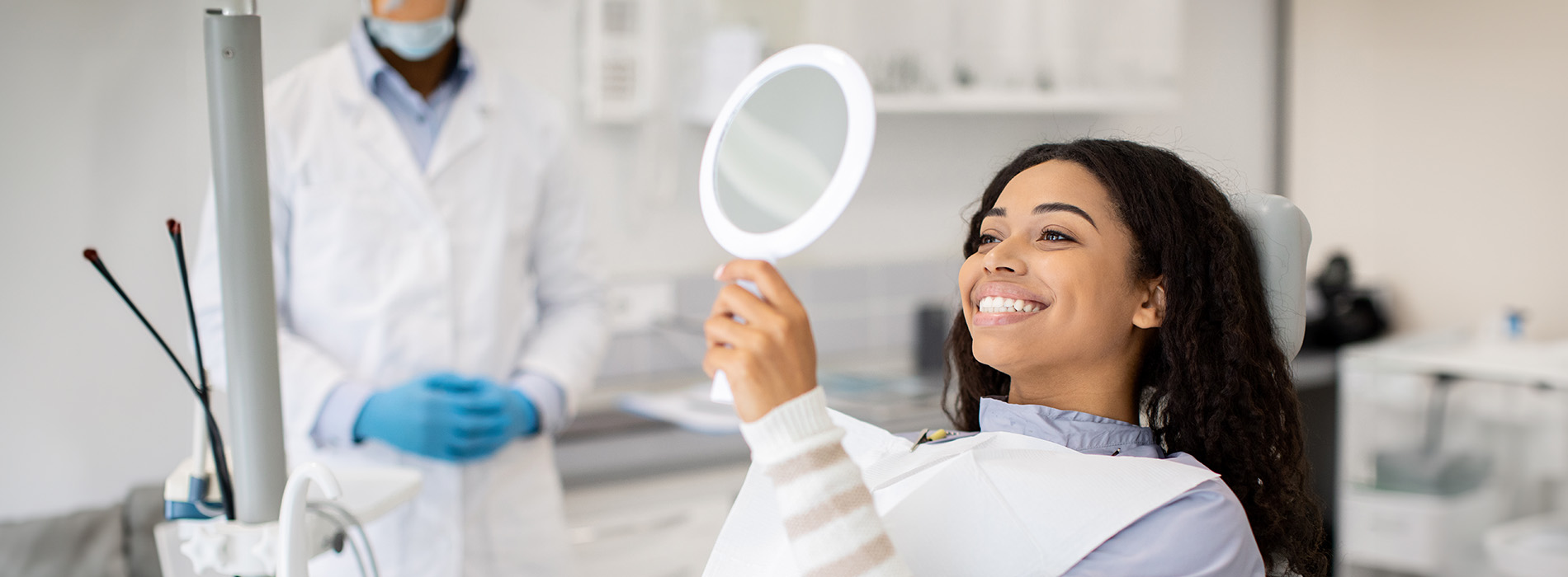 The image depicts a woman with a smile, looking at her reflection in a mirror while standing in a laboratory setting, surrounded by scientific equipment and wearing protective gloves, with another person observing from behind.