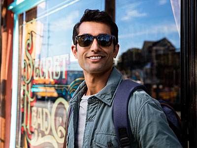 A man wearing sunglasses stands outside a shop with a backpack on his shoulder.