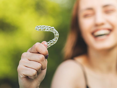 The image shows a person holding a toothbrush with a smiley face on it against a blurred background.