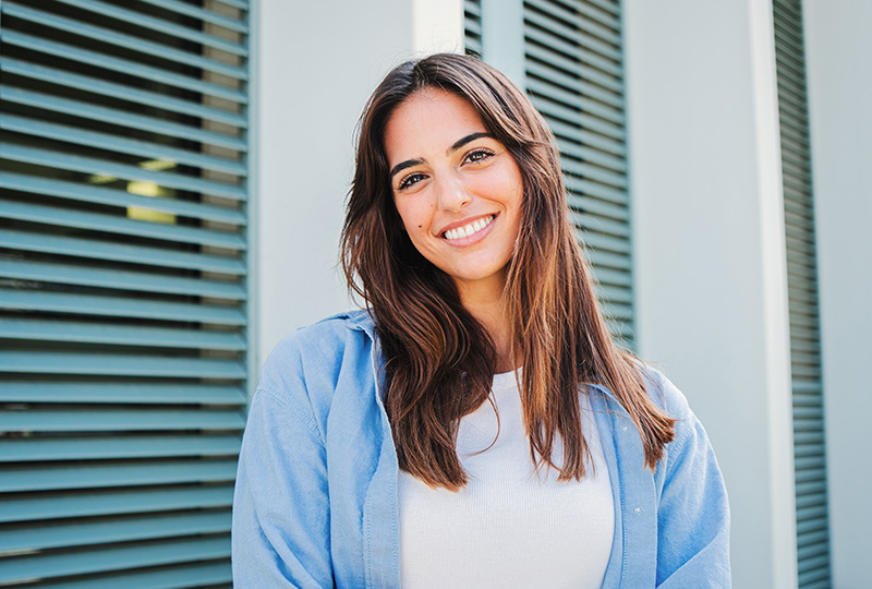A young woman with long hair, wearing a blue top, smiling at the camera while standing against a backdrop of vertical slats.