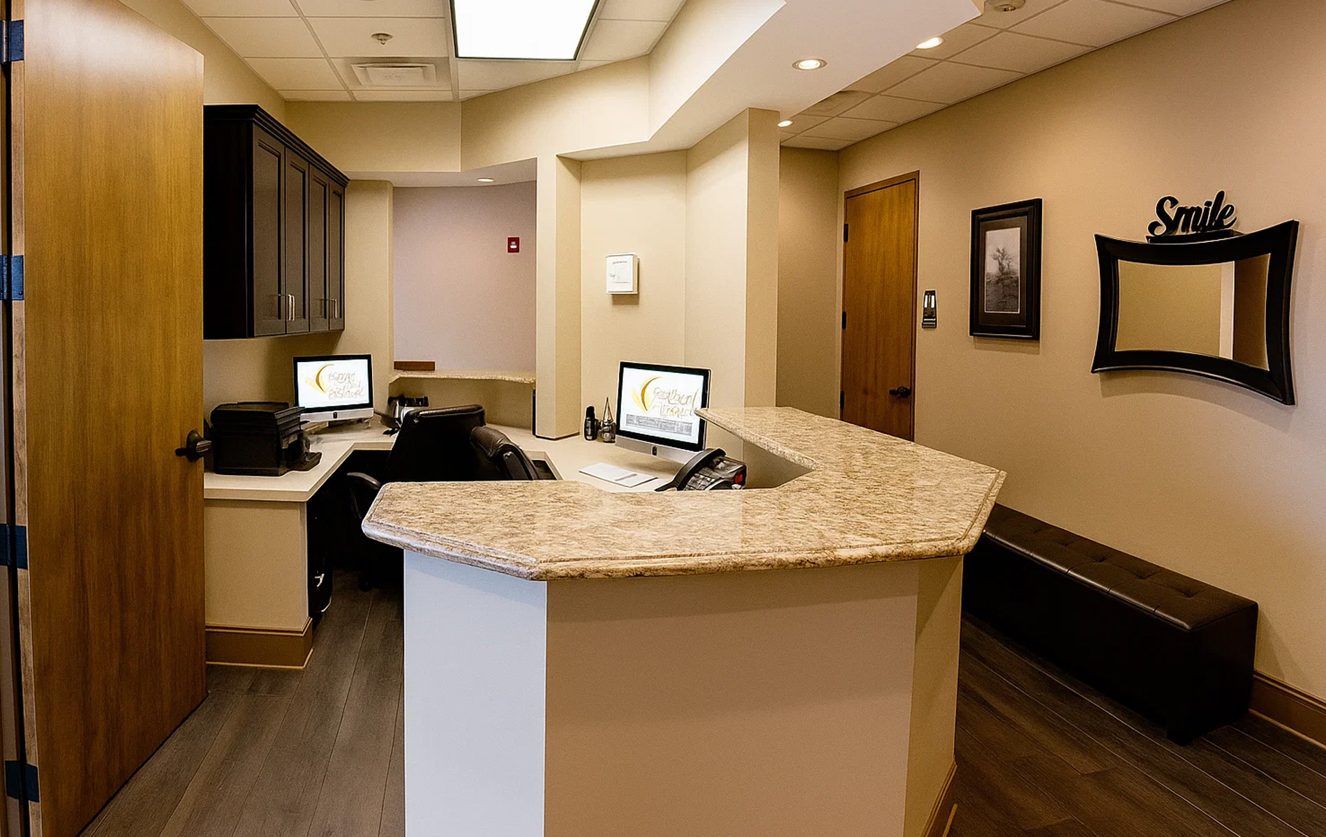 The image shows an interior space that appears to be a medical office reception area with a counter and a computer monitor on the counter, along with chairs for seating. There are framed pictures on the wall and the floor is tiled.