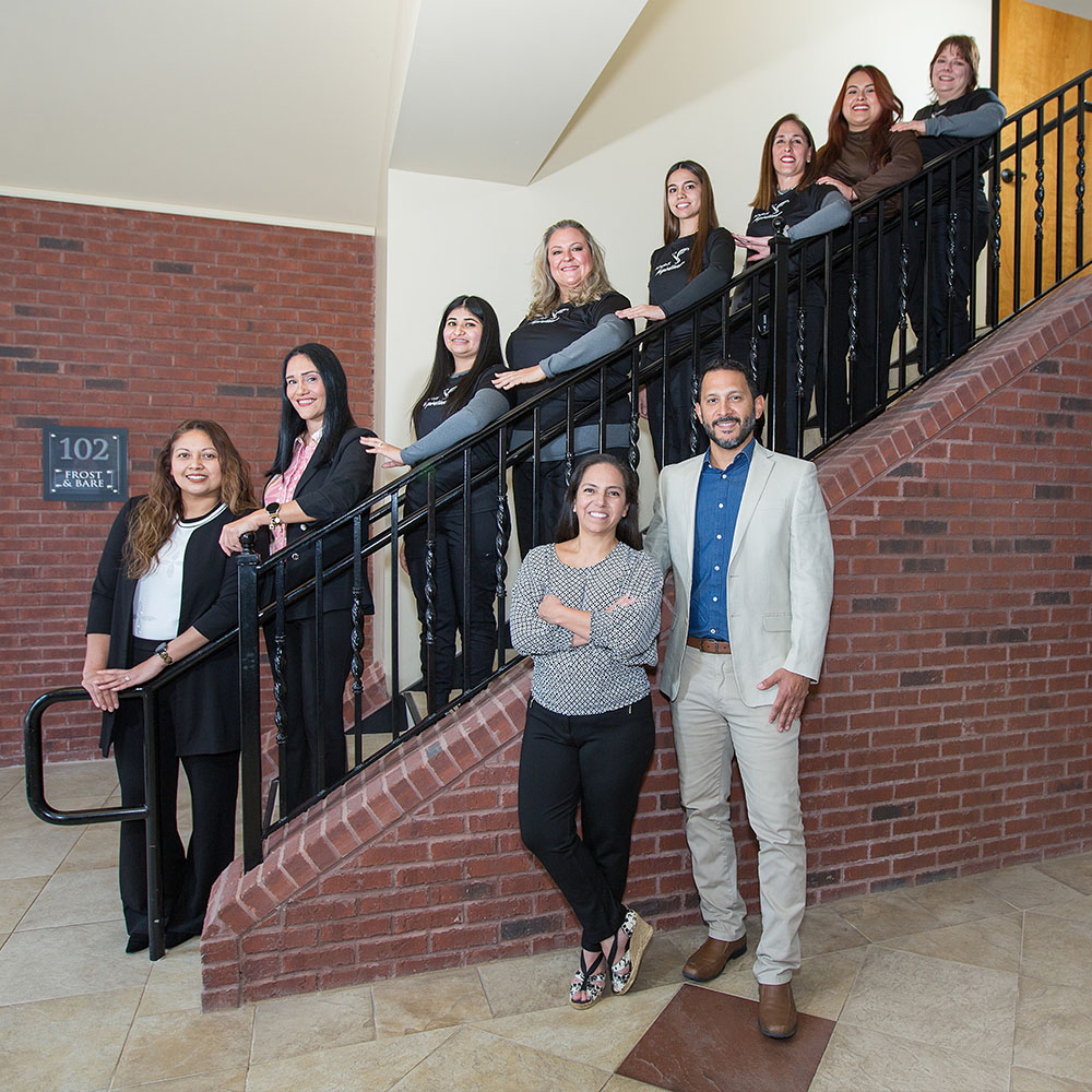 The image shows a group of individuals posing on a staircase with a handrail, including one man standing at the bottom and smiling towards the camera, and several other people standing behind him, some of whom are also smiling and looking directly at the camera, while others appear to be looking at something off-camera. The setting seems to be an indoor environment, possibly a lobby or entrance area, with a brick wall in the background and a sign that reads '101.' There is no visible text providing additional context about the event or the people depicted.