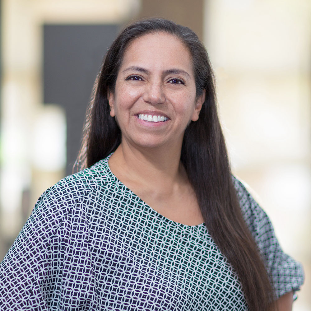The image shows a woman with long hair smiling at the camera. She has a light complexion and is wearing a patterned top. The background appears to be an indoor setting with a blurred image of another person in the distance, suggesting a professional or public environment.