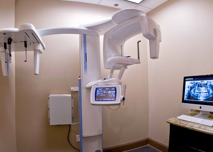 The image shows a dental office with modern equipment, including a dental chair, a large digital X-ray machine, and a computer monitor displaying an X-ray image. The room has a professional appearance with white walls and a beige carpeted floor.