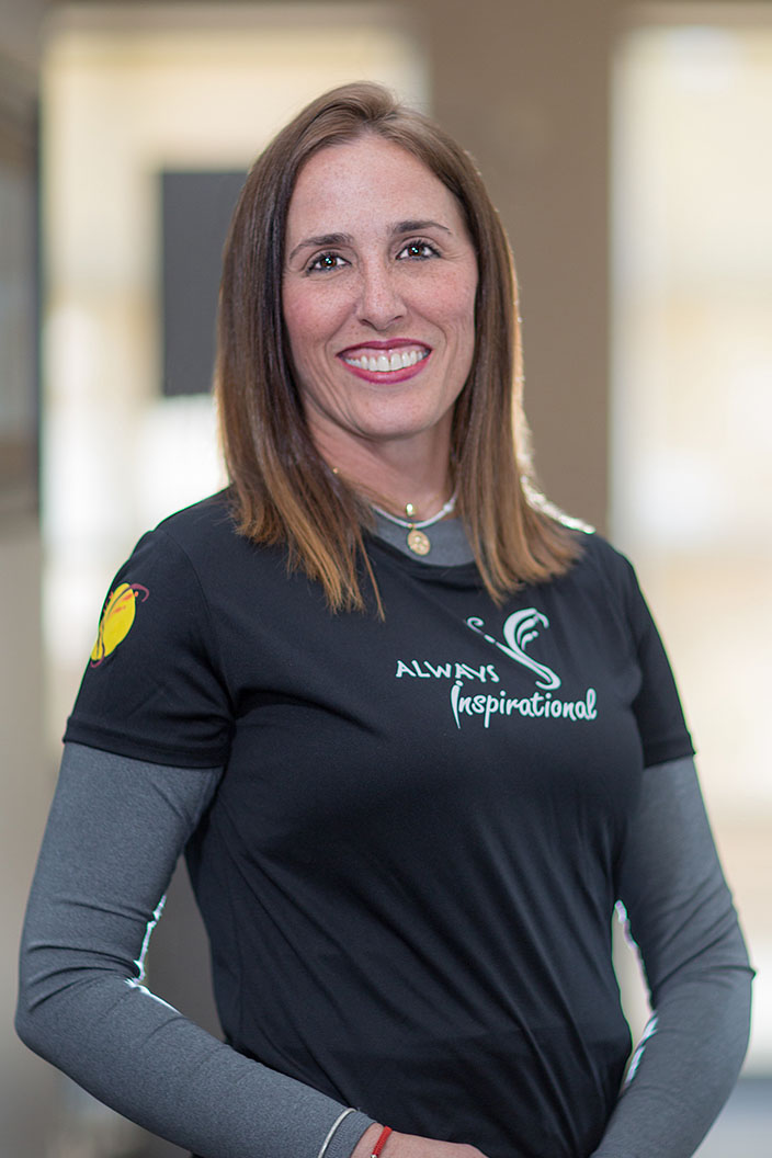 The image shows a woman standing indoors, wearing a black t-shirt with white text and a logo, and a matching black visor. She has short brown hair and is smiling at the camera.