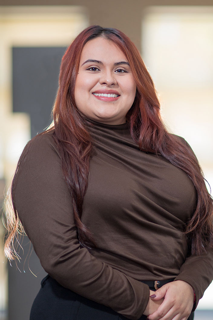 The image shows a smiling woman with long red hair, wearing a brown top, standing against a blurred background.