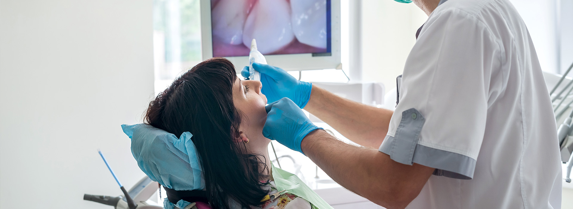 The image shows a dental professional performing a procedure on a patient s mouth, with the patient seated in a dental chair and the professional standing over them using dental instruments.