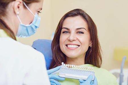 The image shows a woman with a broad smile sitting at a dental chair receiving a dental treatment from a professional wearing medical gloves, likely in an oral health care setting.