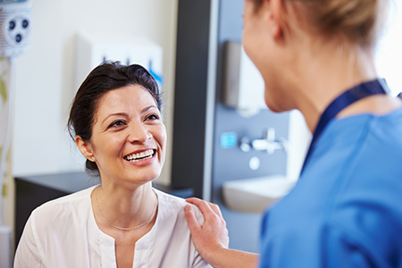 A photograph showing two individuals, likely in a medical setting, with one person seated and smiling at the camera while another stands behind them, both dressed professionally, suggesting a professional interaction between healthcare providers and a patient.