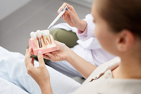 A woman holding a pink tooth model with a dental tool, standing in front of a medical professional who appears to be observing her work.