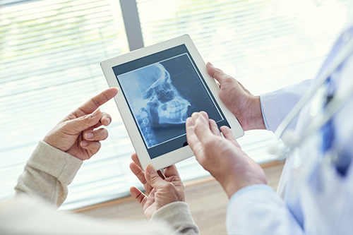 A medical professional is showing a tablet with an X-ray image to two patients in a clinical setting, using a stylus to point at the screen.