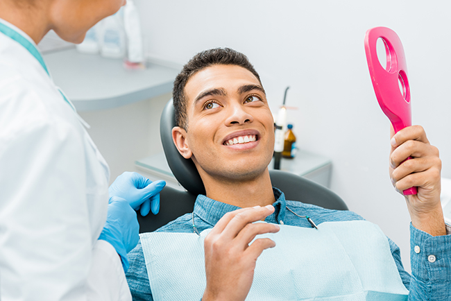 The image shows a person sitting in a dental chair with a smile on their face, holding a pink object that appears to be a dental device, while being attended to by a dental professional wearing a white coat and stethoscope.