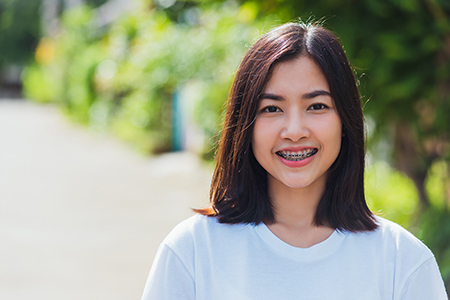The image shows a person with short hair smiling at the camera, wearing a white top, against a blurred outdoor background.