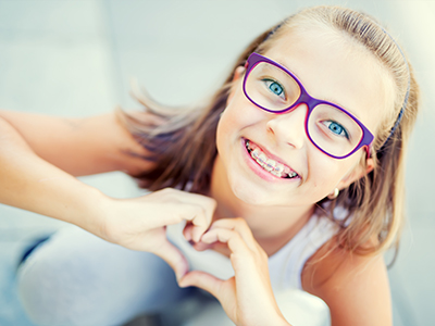 The image depicts a young girl with glasses smiling at the camera while holding her hands together to form a heart shape, with a background that appears to be an outdoor setting.